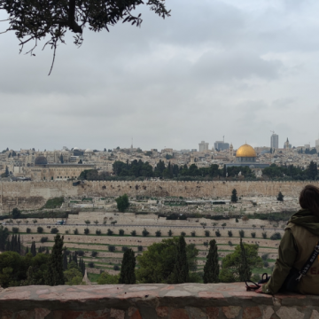 Eappi-tarkkailija istuu kiviaidan reunalla ja katselee Jerusalemin siluettia. Taustalla näkyy Kalliomoskeija.