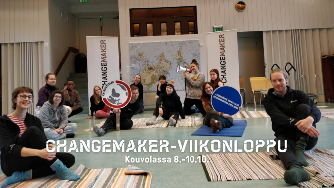 Changemaker-viikonlopun osallistujia, joilla on Changemkaer-kylttejä. Lattialla on räsymattoja ja einällä maailman kartta.