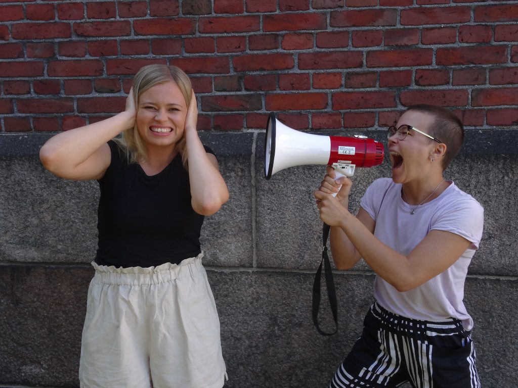 Nuoret poseeraavat tiiliseinän edessä. Toisella on kädessään megafoni jonka hän on kohdistanut toista kohti, joka pitelee korviaan.