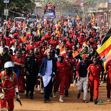 Väkijoukko punaisissa vaatteissa marssii hiekkatietä eteenpäin Ugandan lippu käsissä.