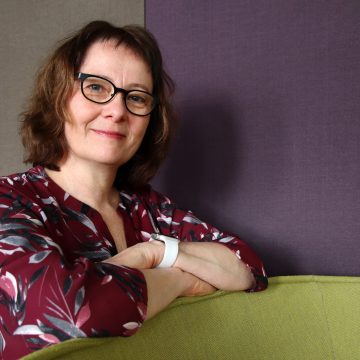 Nainen viininpunaisessa paidassa nojaa huonekalun reunaan kädet ristittynä ja katsoo kohti kameraa.