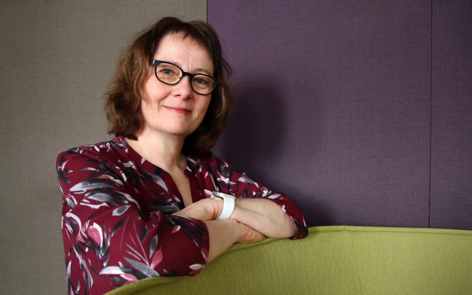 Nainen viininpunaisessa paidassa nojaa huonekalun reunaan kädet ristittynä ja katsoo kohti kameraa.