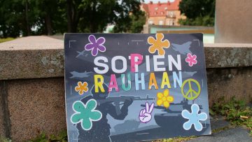 Värikäs kyltti, jossa kukkia ja teksti "Sopien rauhaan" nojaa maassa suihkulähteen reunaan.