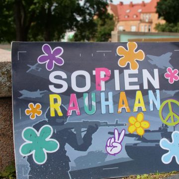 Värikäs kyltti, jossa kukkia ja teksti "Sopien rauhaan" nojaa maassa suihkulähteen reunaan.