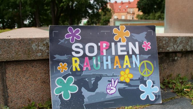 Värikäs kyltti, jossa kukkia ja teksti "Sopien rauhaan" nojaa maassa suihkulähteen reunaan.
