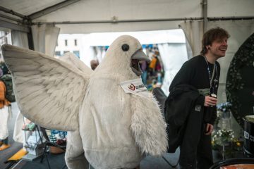 Rauhankyyhky ja mustapaitainen hymyilevä henkilö kohtaavat ihmisiä teltassa.