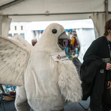 Rauhankyyhky ja mustapaitainen hymyilevä henkilö kohtaavat ihmisiä teltassa.