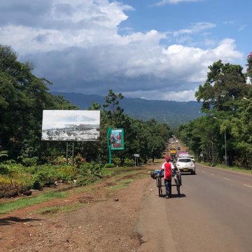Kuvassa tansanialainen maantie, jossa kulkee autoja ja ihminen kärryn kanssa.